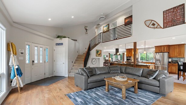 A spacious living area with a gray sectional sofa, a wooden coffee table, and a blue striped rug. A staircase leads to an upper level with a kitchen and dining area.