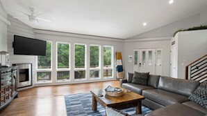 A spacious living room with a large window, a flat-screen TV, a fireplace, and a staircase.