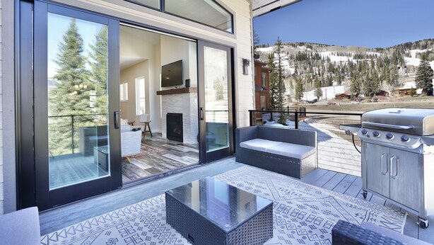 A modern outdoor deck with a glass door leading to a living area, a barbecue grill, and a view of a snowy mountain landscape. - Hot Tub, Modern Home, Hiking and Fishing Close! (Durango)