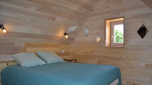 Dormitorio de cabaña de madera con una cama, dos almohadas, una mesita y una ventana con vistas a la vegetación.