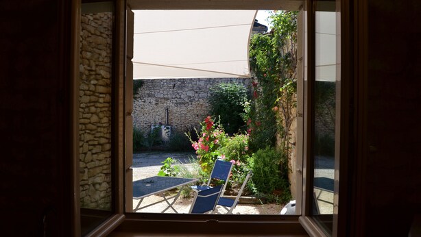 Vista a través de una ventana: un muro de piedra, un jardín con mesa y sillas, y un edificio al fondo.
