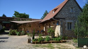 Una casa de piedra con techo inclinado, un porche cubierto con mobiliario exterior y un jardín con diversas plantas y flores.