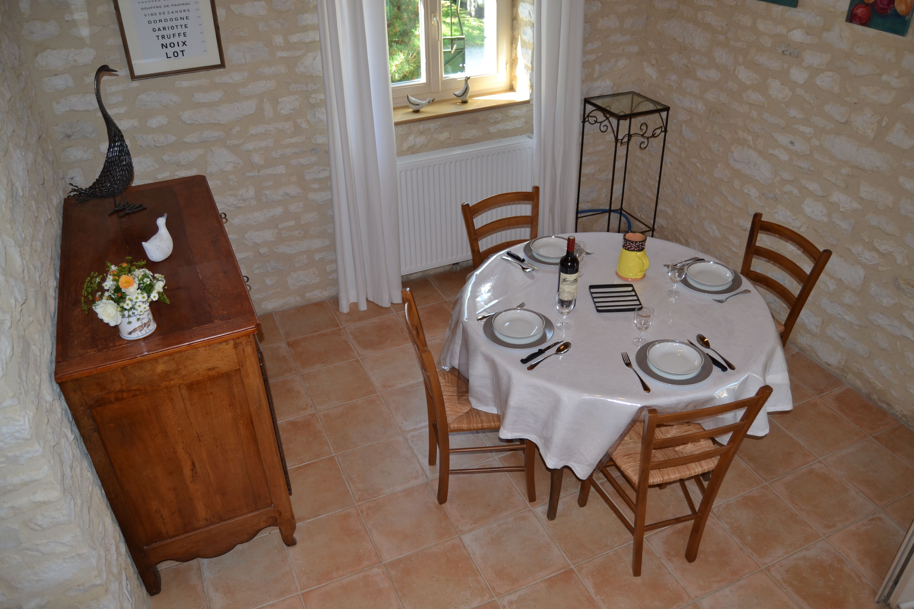 A dining area with a round table set for four, surrounded by chairs. A wooden cabinet with a vase of flowers is to the left.