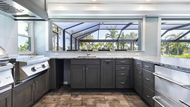 A modern kitchen with dark cabinetry, stainless steel appliances, and a large window overlooking a tropical landscape. - 🌺Urlaub Haus | A Lakefront Paradise Reimagined in Murex Lakes🐬 (Sanibel)
