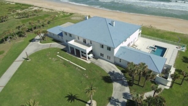 An aerial view of a large, single-story house with a blue roof, situated on a grassy area with a view of the ocean.