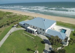 Une vue aérienne d’une grande maison de plain-pied avec un toit bleu, située dans une zone verdoyante et offrant une vue sur l’océan.
