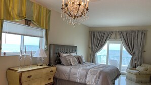 A bedroom with a large bed, a dresser, a chandelier, and a view of the sea.