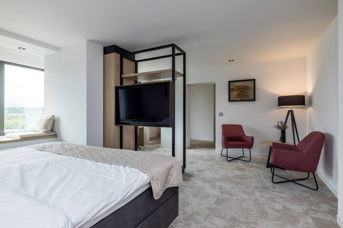 Un dormitorio moderno con cama, televisor montado en un marco negro, una silla roja y un escritorio con lámpara.