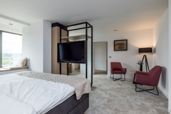 Ein modernes Schlafzimmer mit einem Bett, einem an einer schwarzen Konstruktion befestigten Fernseher, einem roten Sessel und einem Schreibtisch mit einer Lampe.