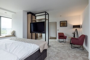 Un dormitorio moderno con cama, televisor montado en un marco negro, una silla roja y un escritorio con lámpara.