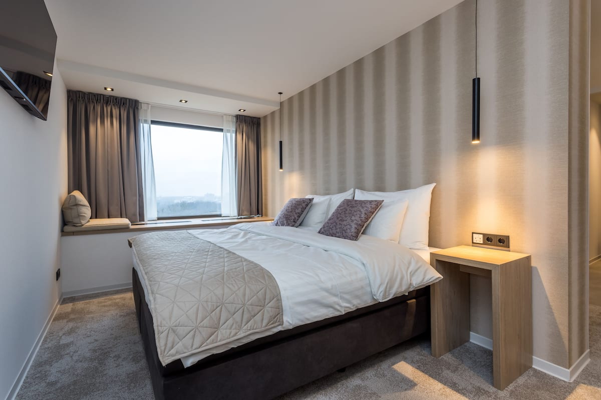 Un dormitorio moderno con una cama grande, una mesita de noche, una ventana con cortinas y una lámpara fijada a la pared.