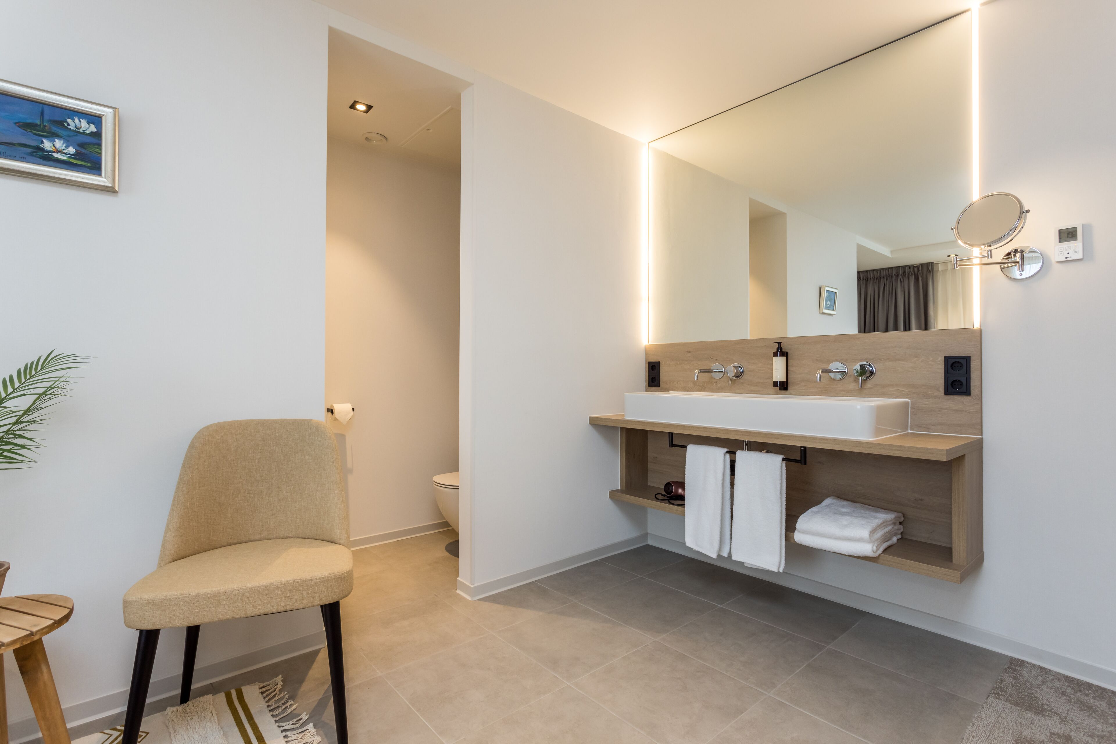 A modern bathroom with a beige chair, a wooden vanity with a sink, and a large mirror.