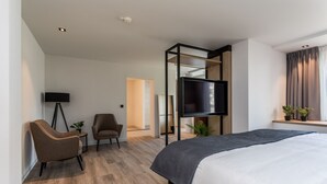 Una habitación de hotel moderna con una cama grande, un televisor de pantalla plana en la pared, una zona de estar con una silla y una mesita, y una ventana con cortinas.