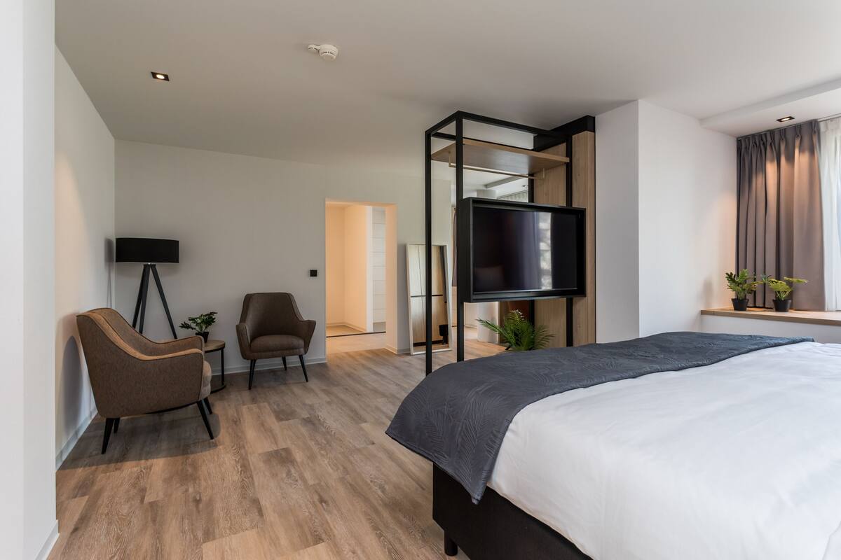 Una habitación de hotel moderna con una cama grande, un televisor de pantalla plana en la pared, una zona de estar con una silla y una mesita, y una ventana con cortinas.