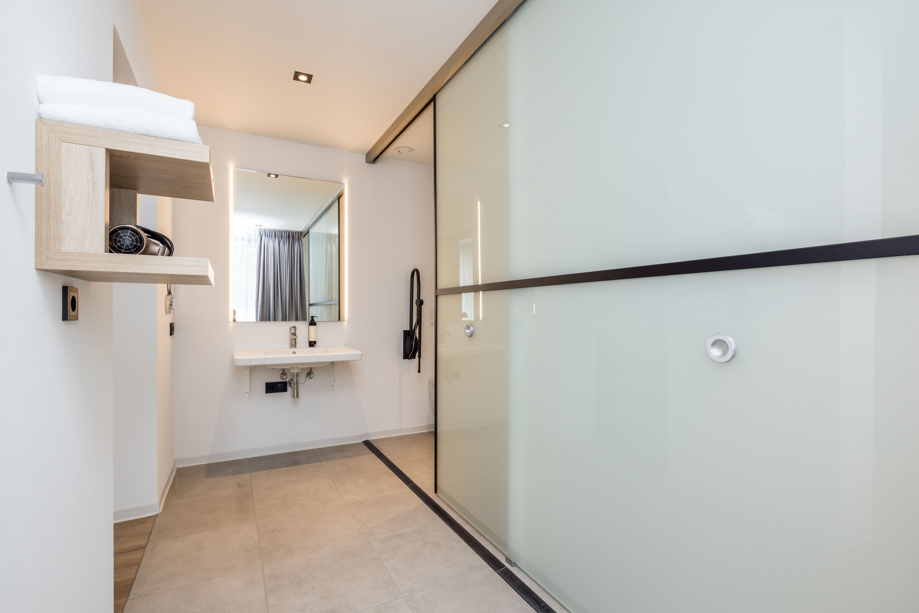Un baño moderno con cabina de ducha de vidrio, estante de madera con artículos de aseo, lavamanos y espejo.