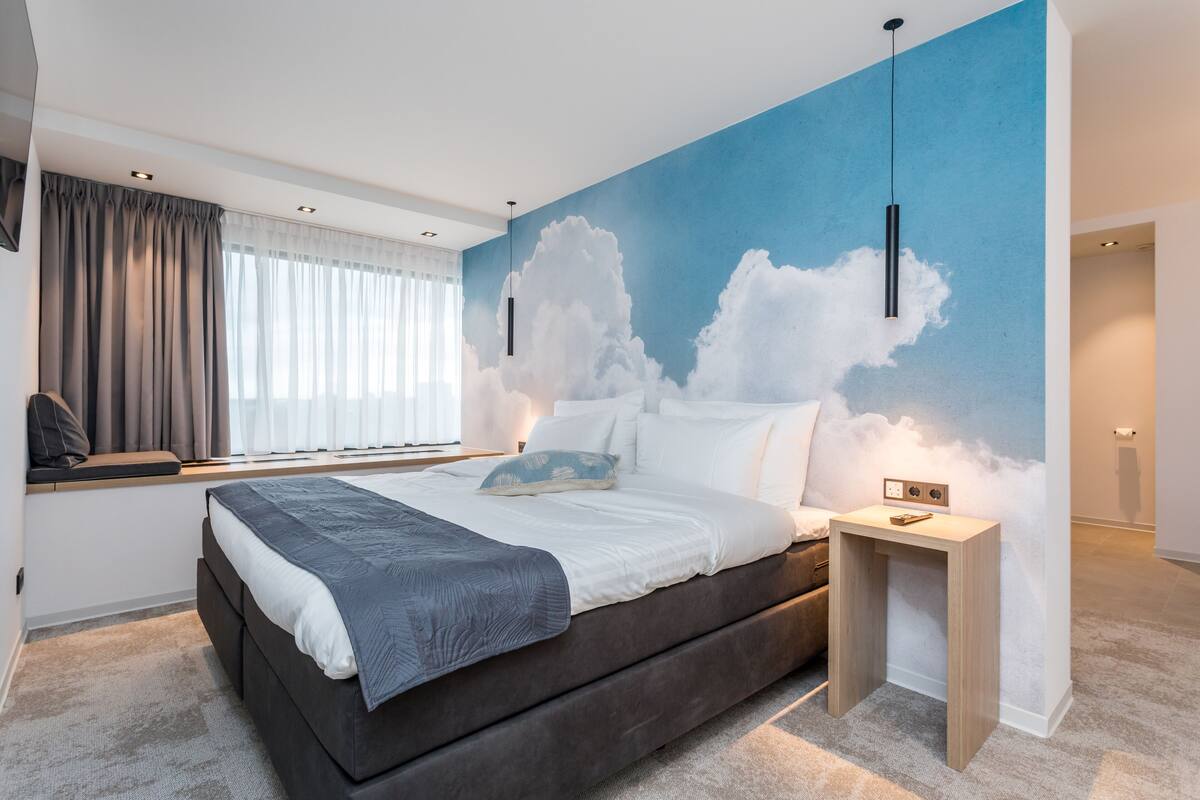 Un dormitorio moderno con una cama grande, una mesita de noche, un mural en la pared con nubes y una ventana con cortinas.