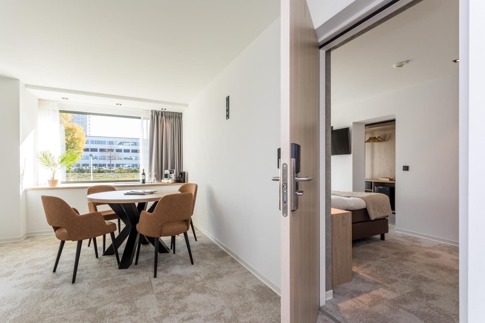 A modern hotel room with a dining area, a bed, and a view of the city.