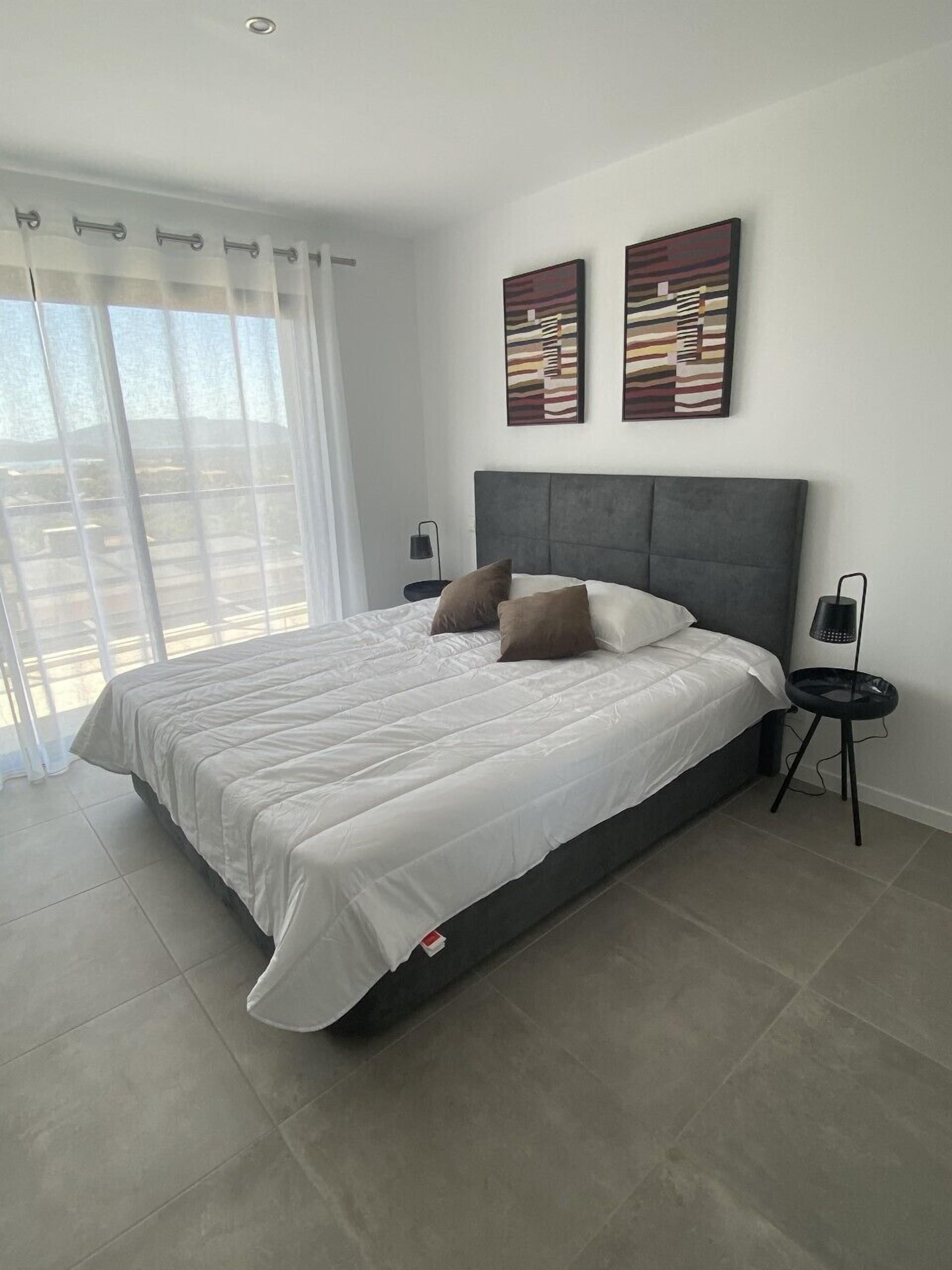 A modern bedroom with a large bed, two wall-mounted artworks, a bedside table with a lamp, and a view through a large window.