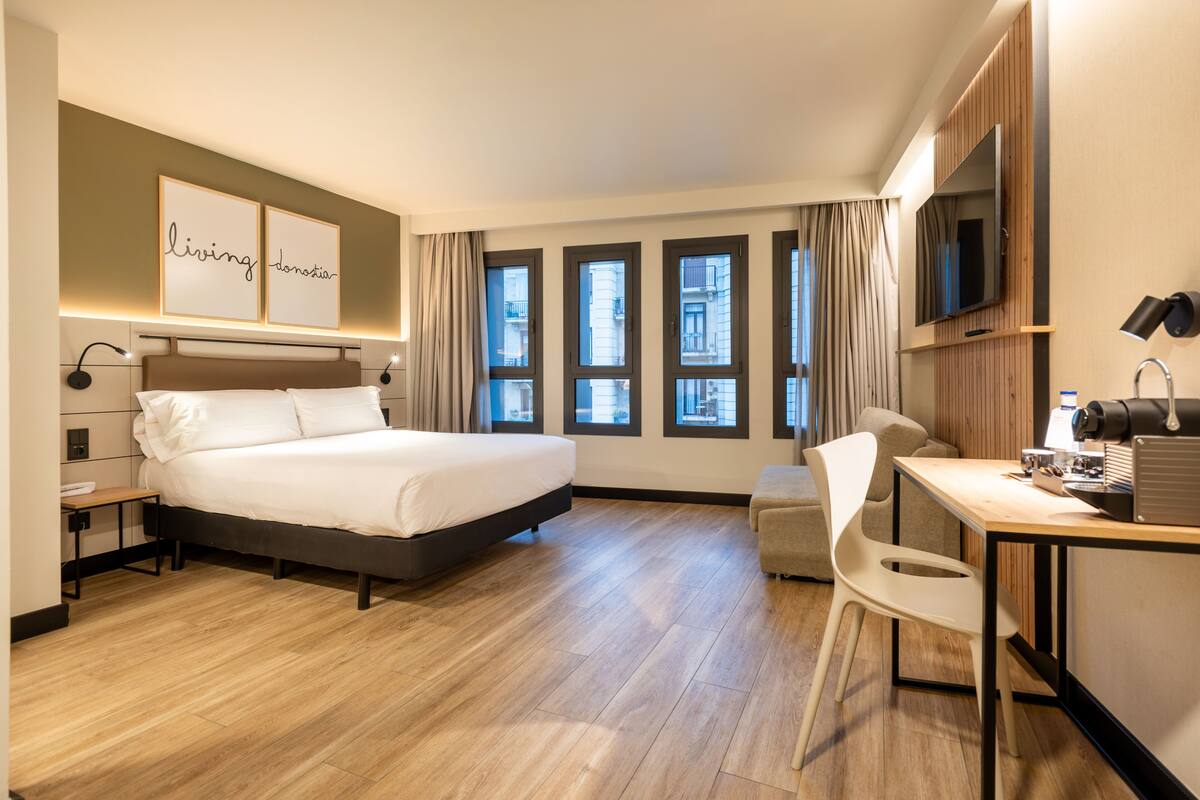 Een moderne hotelkamer met een groot bed, een bureau met stoel, een flatscreen televisie en meerdere ramen met uitzicht over de stad.
