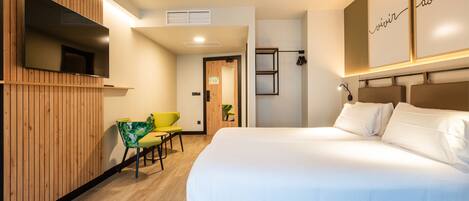 A modern hotel room with a large bed, wooden paneling on one wall, a flat-screen TV, and a small seating area with a chair and table.