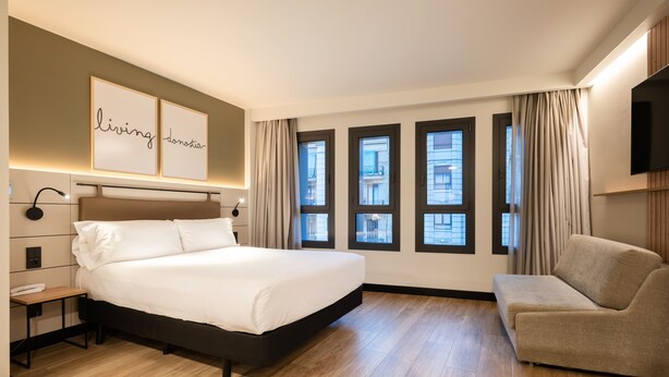 A hotel room with a large bed, a bedside table, a chair, a TV, and large windows with curtains. - Leonardo Boutique Hotel San Sebastián (San Sebastián)