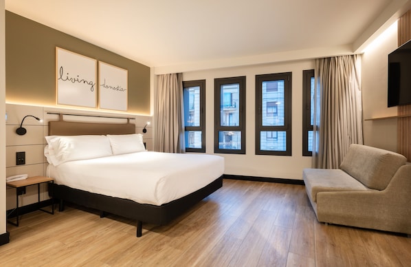 Habitación de hotel con una cama grande, una mesita de noche, una silla, un televisor y ventanas amplias con cortinas.
