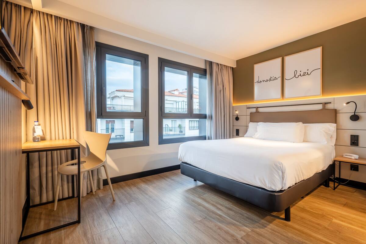 Een moderne hotelkamer met een groot bed, een bureau en twee ramen met uitzicht op gebouwen.