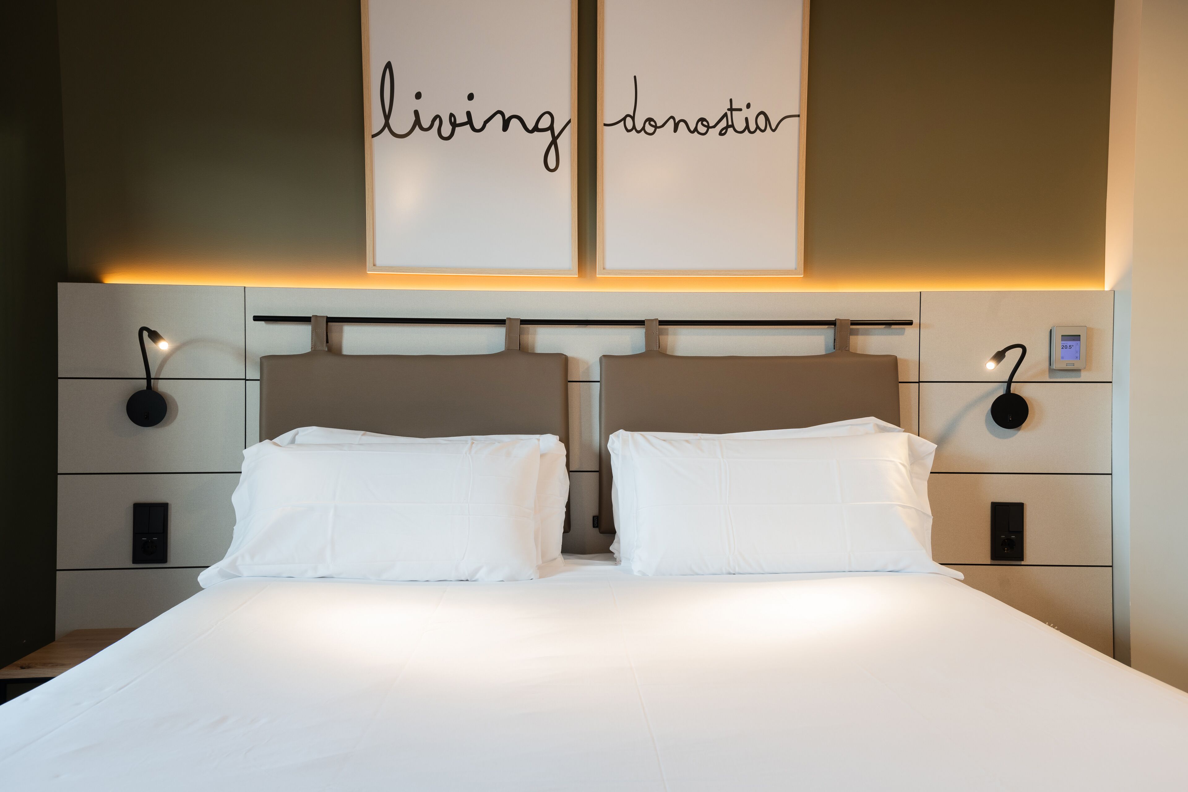 Une chambre d’hôtel avec un grand lit, deux tables de chevet avec des lampes, un panneau de commande fixé au mur et une pancarte au-dessus du lit sur laquelle est écrit « living donostia ».