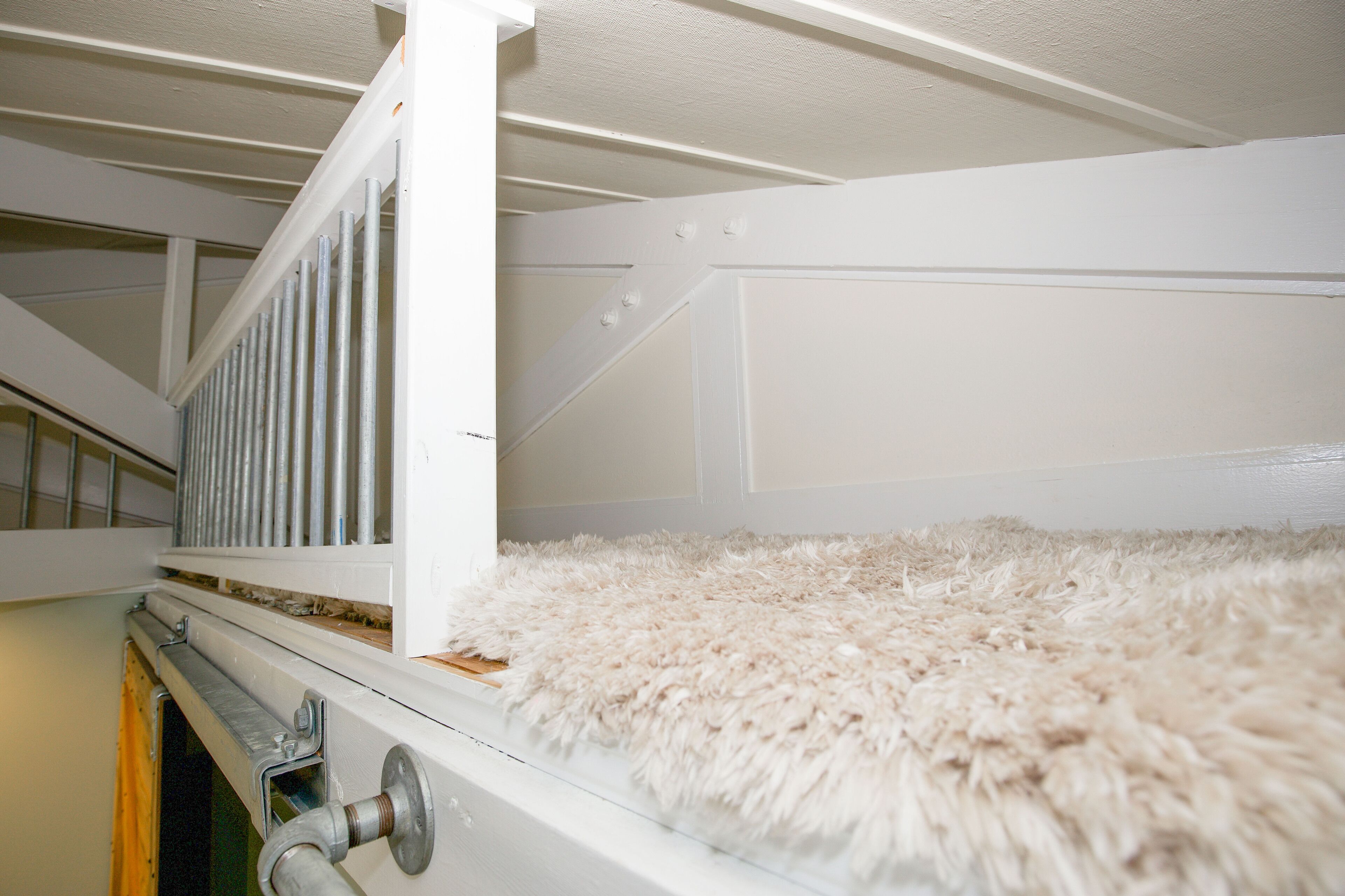 Una stanza con soffitto alto e bianco, tappeto bianco e soffice, ringhiera in metallo e pavimento in legno.