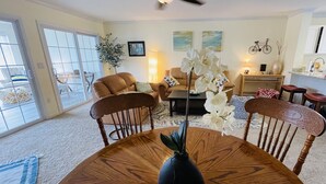 A spacious living room with a ceiling fan, a dining table with chairs, a coffee table, and a large window with a view. - Renovated & Spacious Retreat on East Rt 1 – Pool, Bike Trails, Mins to Beach! (Lewes)