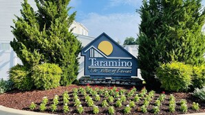 A sign for Taramino The Villas of Lewes with a house-shaped logo, surrounded by greenery and a flower bed. - Renovated & Spacious Retreat on East Rt 1 – Pool, Bike Trails, Mins to Beach! (Lewes)