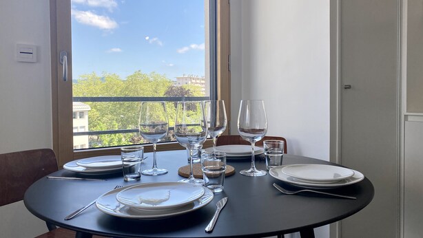 Mesa de comedor dispuesta con platos, cubiertos y copas de vino, ubicada cerca de una ventana con vistas a edificios y árboles.