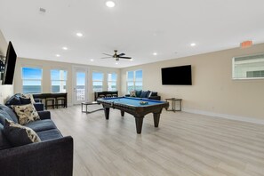 A spacious living room with a pool table, a sofa set, and a large window offering a view of the ocean.