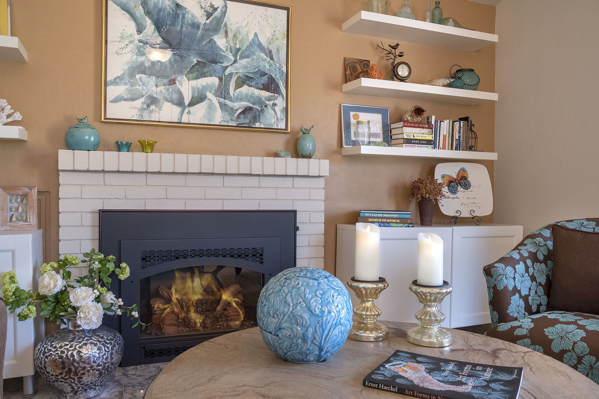 A living room with a fireplace, a blue decorative vase, two candles, and a book titled 'Art Forms in Nature' on a table.