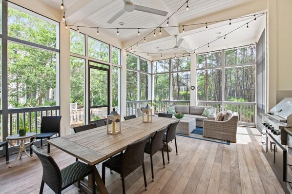 A spacious patio with a wooden table and chairs, large windows offering views of trees, and ceiling fans.