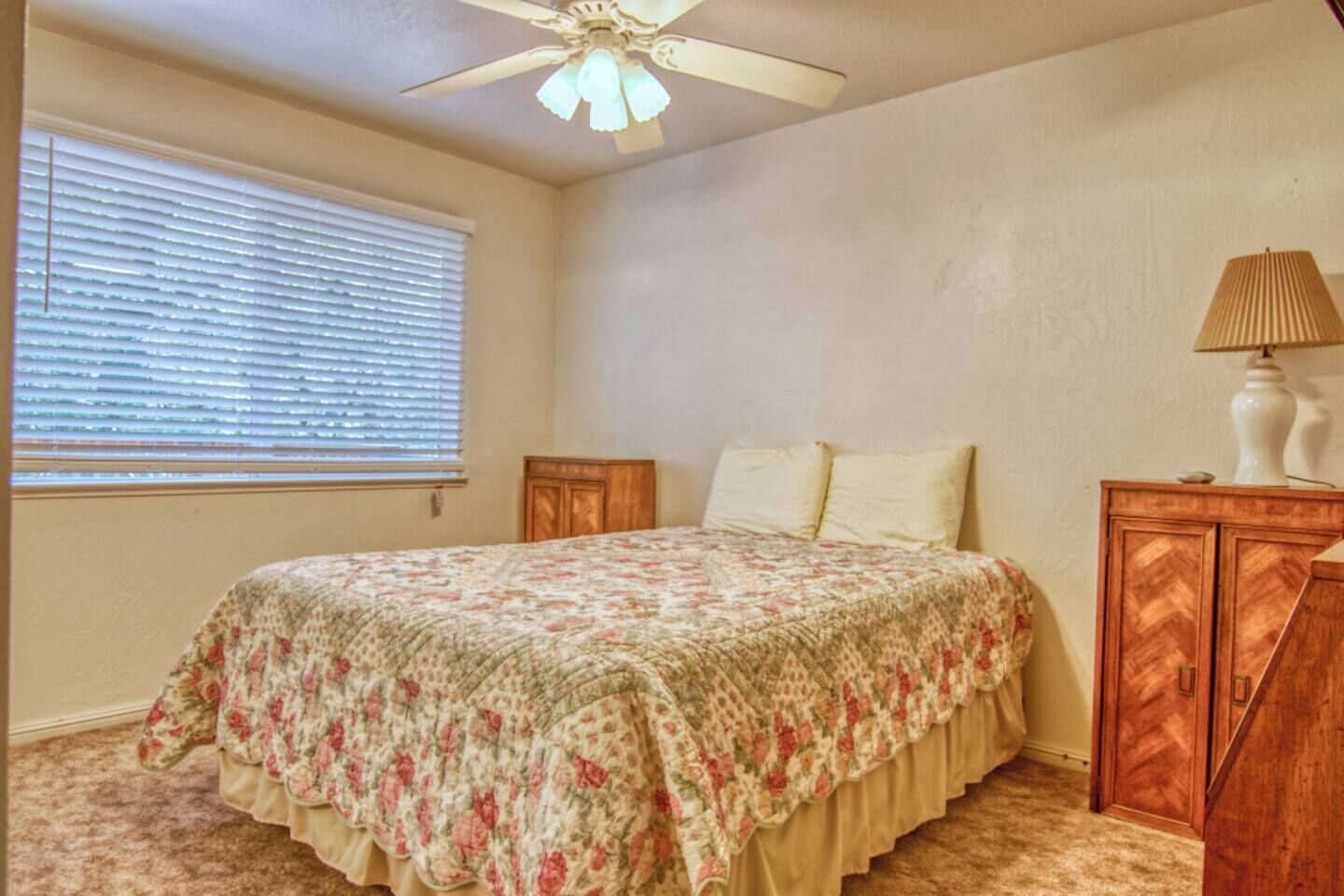 Une chambre à coucher comprenant un lit, un ventilateur de plafond, une fenêtre avec des stores et une table de chevet avec une lampe.