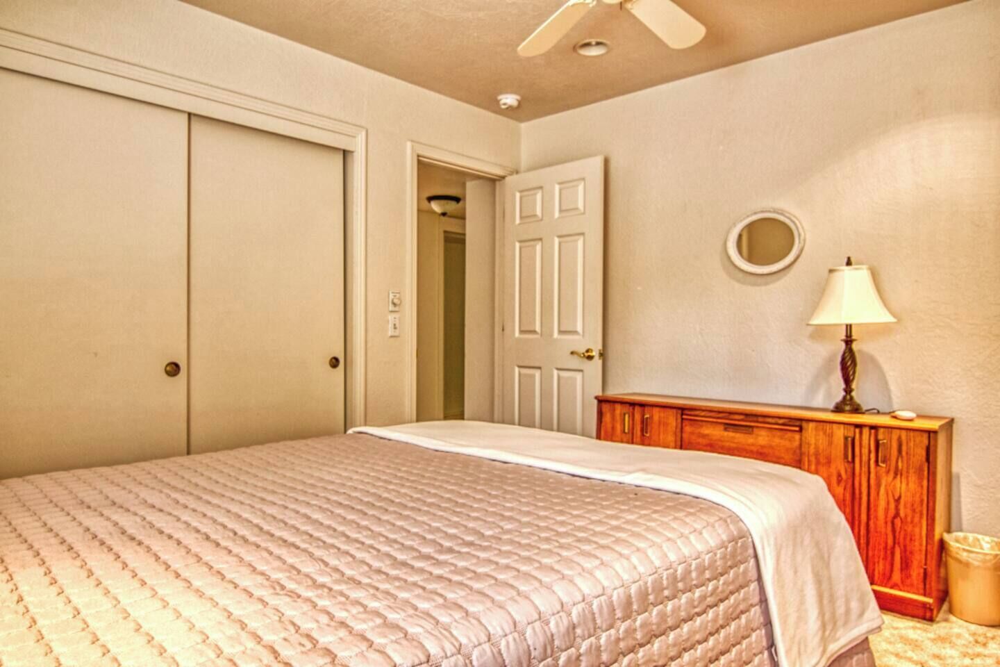 A bedroom with a bed, a wooden dresser, a ceiling fan, and a door leading to another room.