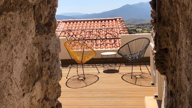An outdoor terrace with a view of mountains, featuring a rustic stone archway, a tiled roof, and two wireframe chairs. - Charmante Maison Avec Toit Terrasse (Montalba-le-Château)