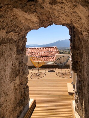 An outdoor terrace with a view of mountains, featuring a rustic stone archway, a tiled roof, and two wireframe chairs.