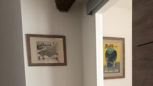 A narrow hallway with white walls, a framed map, a poster, and a wicker bench with a pink figure. - Charmante Maison Avec Toit Terrasse (Montalba-le-Château)