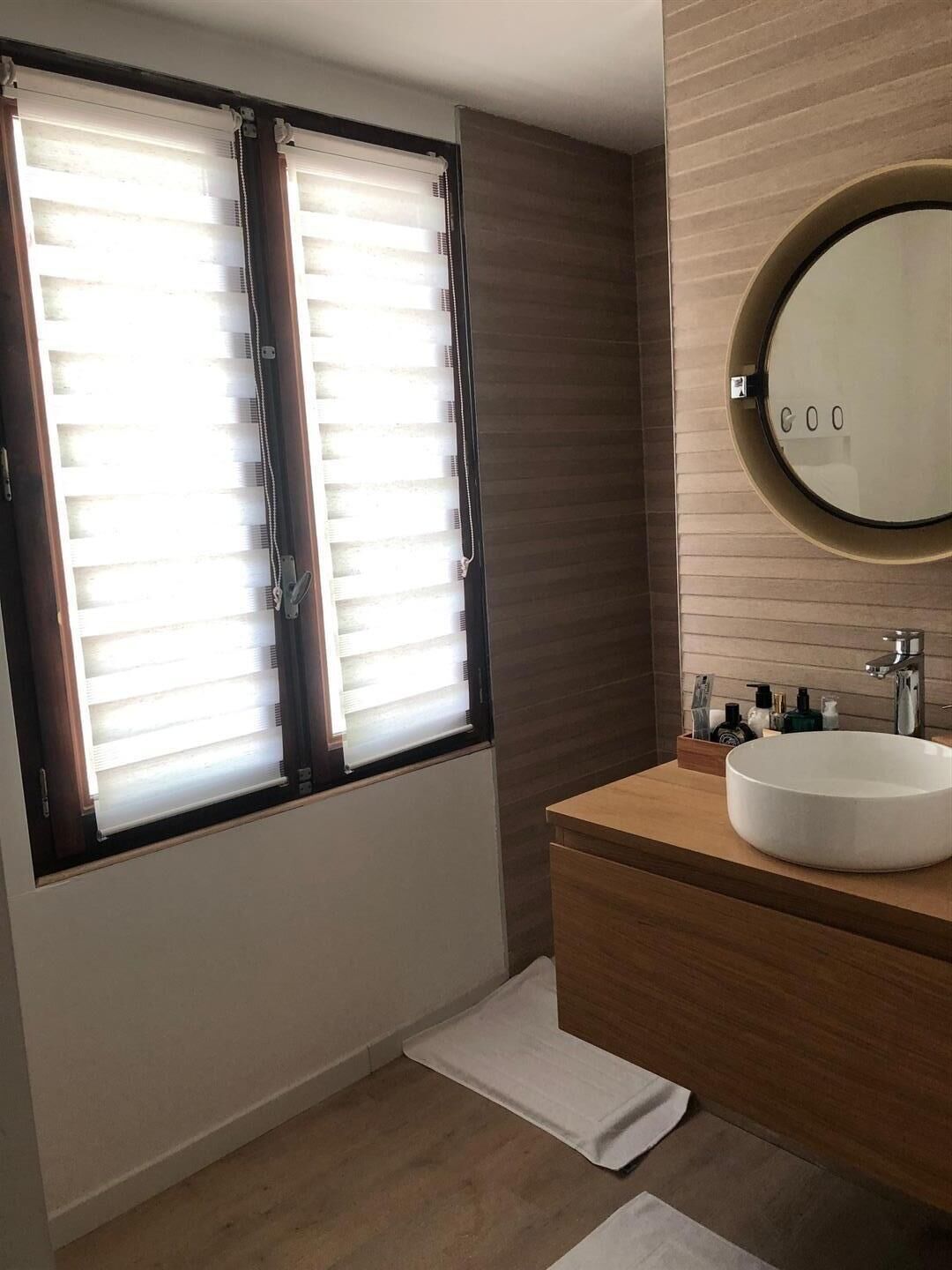 Een moderne badkamer met een groot raam, een ronde spiegel en een houten wastafelmeubel.