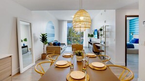 Un espace de salle à manger moderne avec une longue table dressée pour six personnes, un luminaire suspendu et une vue sur l’extérieur.