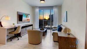 A modern living room with a desk, comfortable seating, a large window with a sunset view, and a flat-screen TV on the wall. - Ocean View| Balcony|Rooftop Pool|Hollywood Beach (Hollywood)