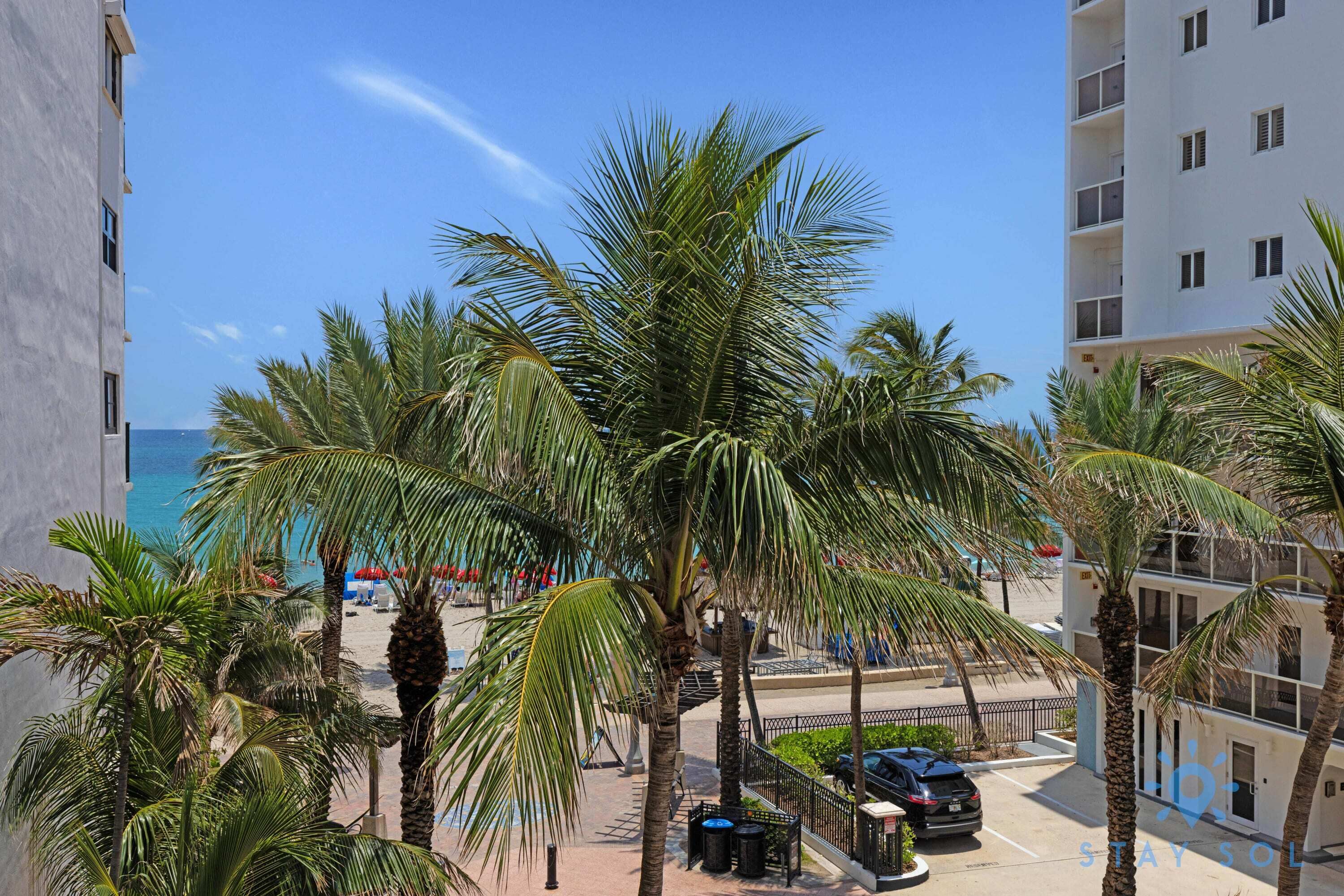 Een hotel aan het strand met palmbomen, een parkeerplaats en uitzicht op de oceaan.