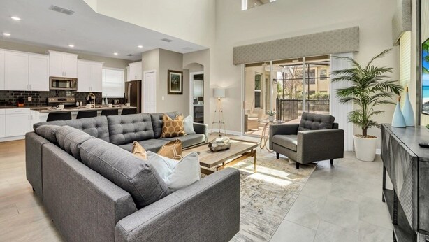 A spacious living area with a large gray sectional sofa, a coffee table, and a fireplace. There is a kitchen area with white cabinets and a microwave. The room has a high ceiling and large windows. - "boutique" Style Home Near Disney! 5 Bedroom Home by RedAwning (Kissimmee)