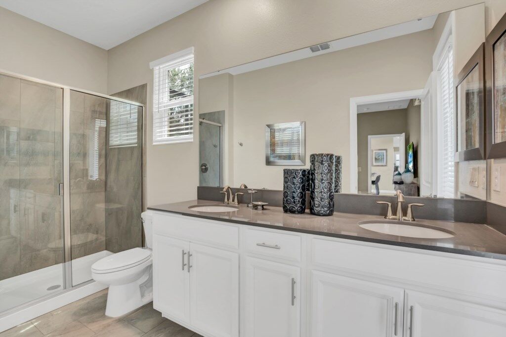 A bathroom with a glass shower enclosure, a toilet, a double sink vanity with two faucets, and a large mirror above the vanity.