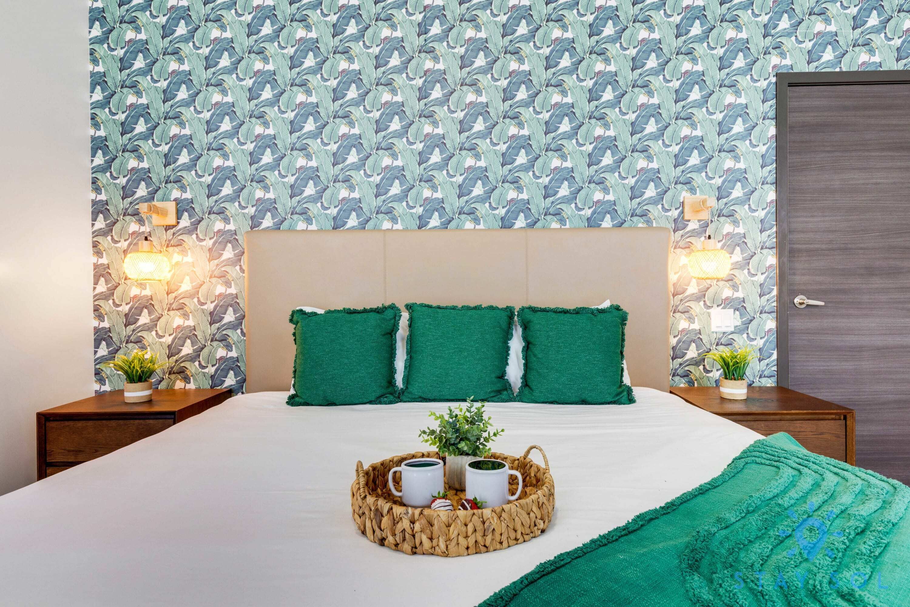 A neatly made bed with a green throw blanket, a tray with two mugs and a plant, and a patterned wallpaper.