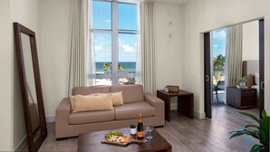 Uma sala de estar com um sofá bege, uma mesa de centro de madeira com uma garrafa de champanhe, copos e um prato de frutas. Há uma janela grande com cortinas, vista para o mar e uma porta que dá para uma varanda com uma cadeira e uma planta.