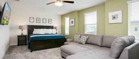 A bedroom with a bed, a gray sofa, a ceiling fan, and framed pictures on the walls.