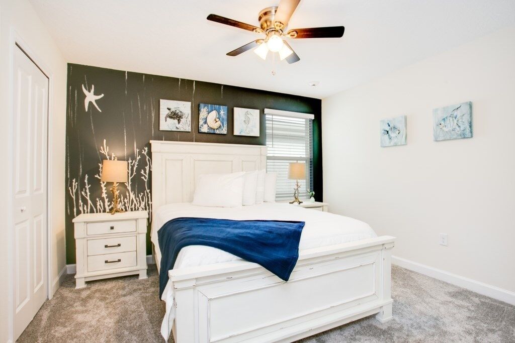 A bedroom with a white bed, a ceiling fan, a nightstand with a lamp, and wall art.